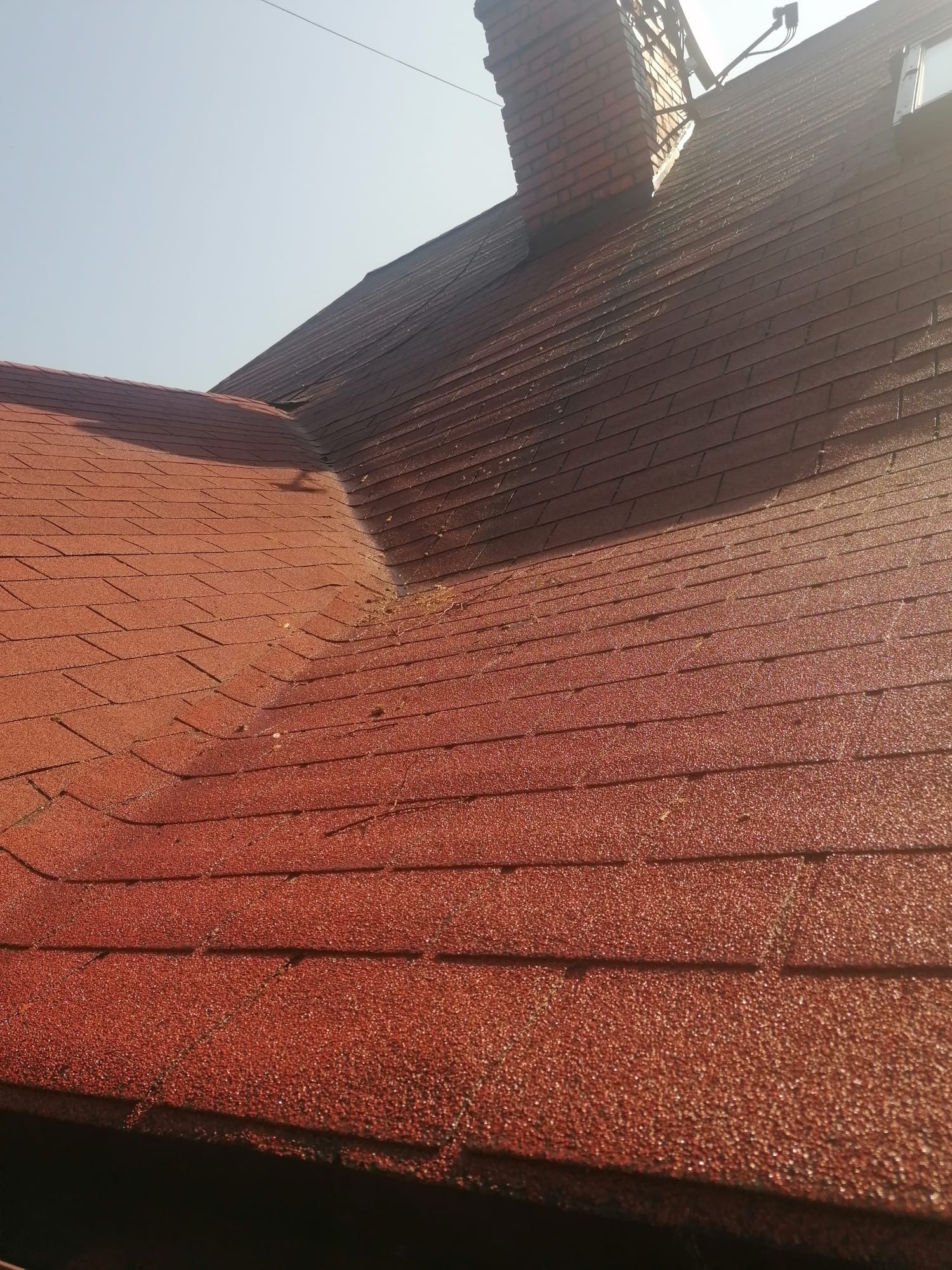 Czerwony dach pokryty gontem bitumicznym, widoczny komin z cegły i fragment zabrudzeń. Ujęcie z dołu, w słoneczny dzień.