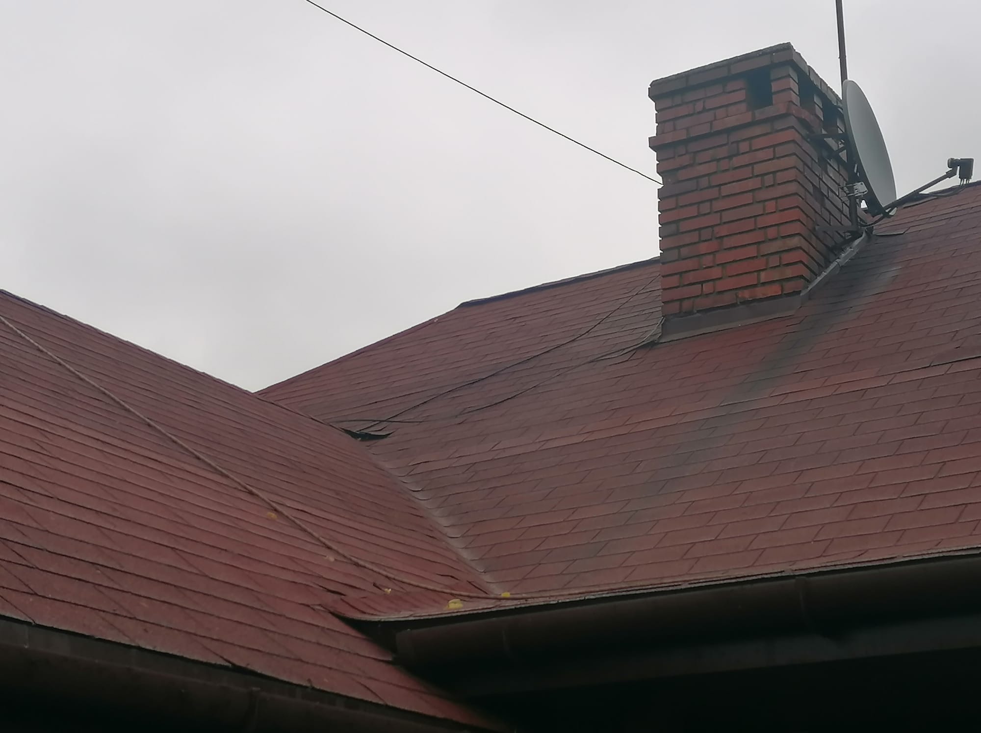 Czerwony dach z gontu bitumicznego z ceglanym kominem i anteną satelitarną na tle pochmurnego nieba. Widoczne zabrudzenia na dachu.