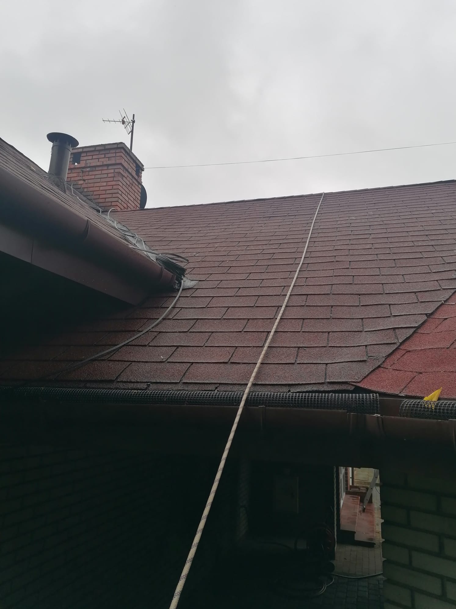 Ciemnobrązowy dach pokryty gontem bitumicznym, z widocznym kominem z cegły i anteną. Wzdłuż dachu biegnie lina zabezpieczająca, a rynna jest zabezpieczona siatką.