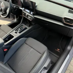 Wnętrze samochodu Cupra: czarna tapicerka fotela z subtelnym wzorem, deska rozdzielcza z ekranem dotykowym i detalami w kolorze miedzianym, eleganckie dywaniki z logo marki.