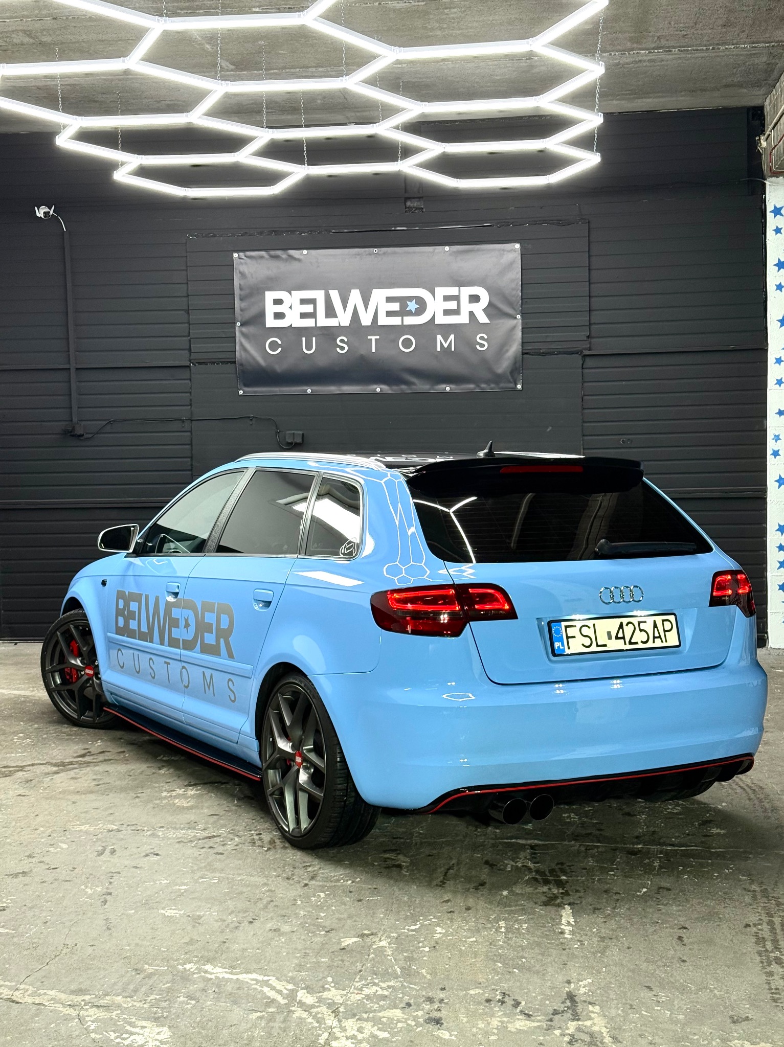 Błękitne Audi A3 po zmianie koloru folią Hexis, z czarnymi akcentami i logo firmy na boku, prezentowane w studiu detailingowym z oświetleniem LED.
