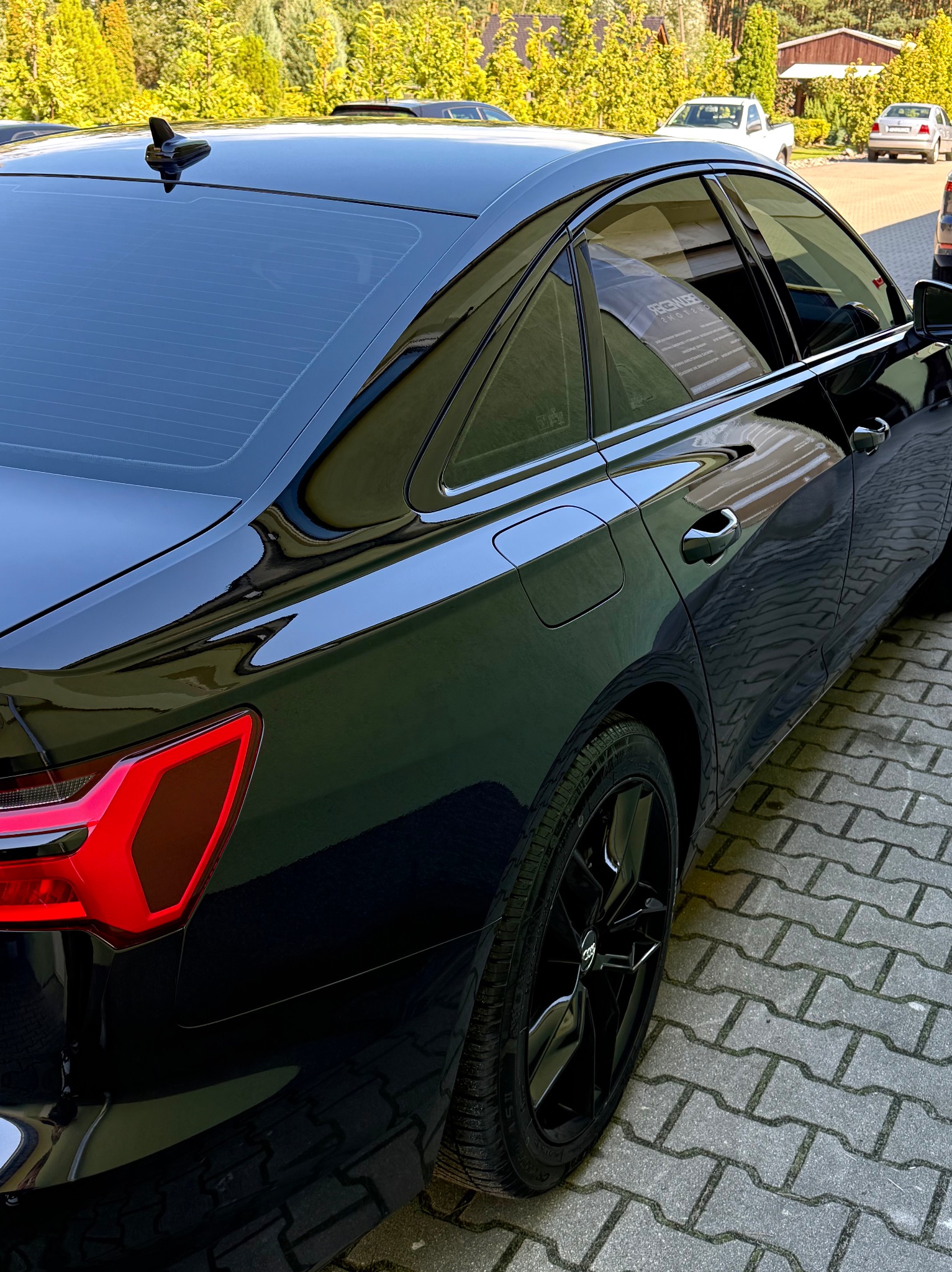 Czarny Audi A6 C8 po dechromingu listw bocznych, z widocznymi detalami karoserii i czarnymi felgami na tle kostki brukowej i zieleni.