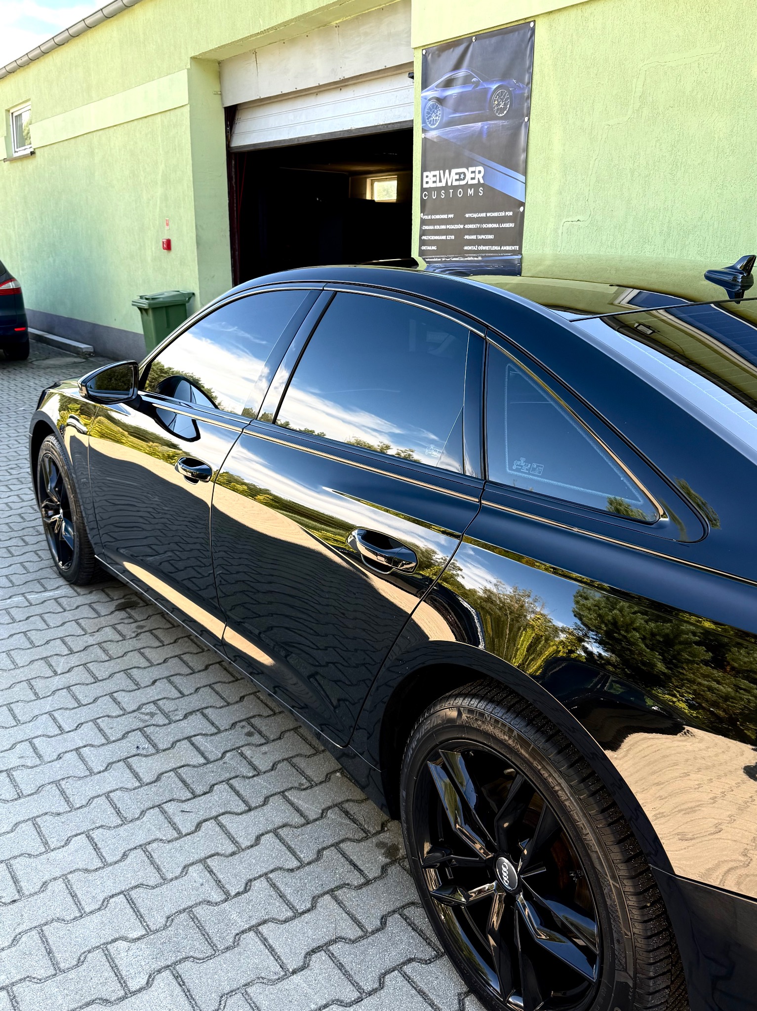 Czarne Audi A6 C8 po dechromingu listw bocznych, z przyciemnionymi szybami, zaparkowane na kostce brukowej przed budynkiem z reklamą usług detailingowych.