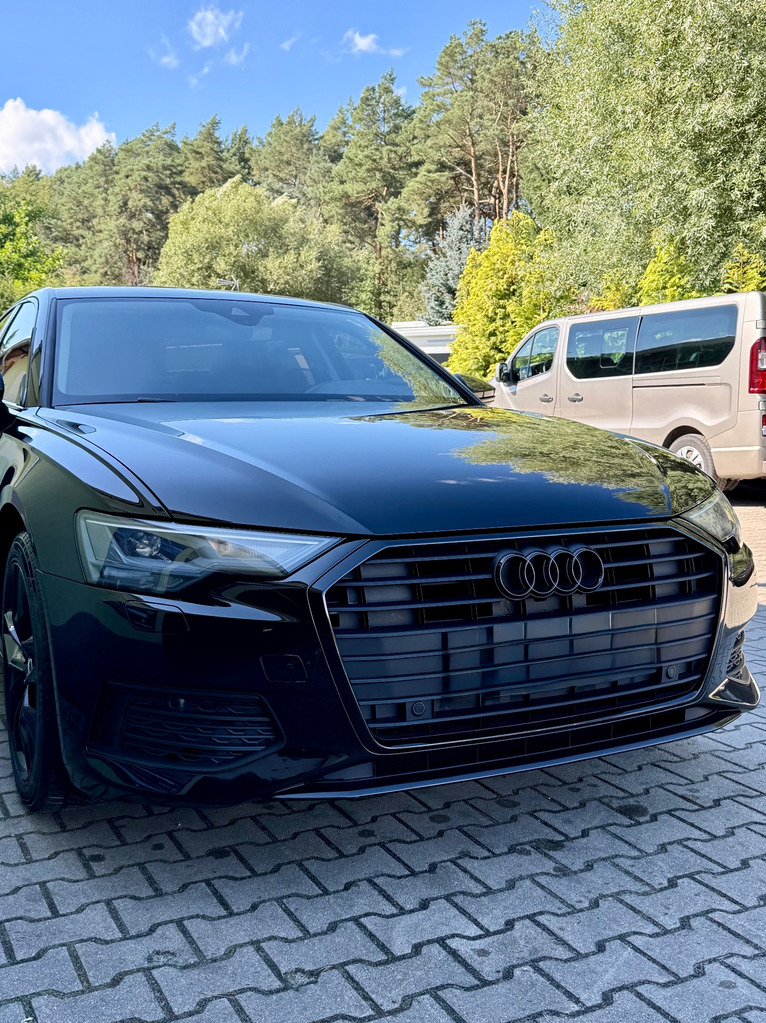 Czarny, lśniący grill Audi A6 C8 po dechromingu, prezentowany z bliska na tle zieleni i beżowego vana. Widoczne detale reflektorów i logo.
