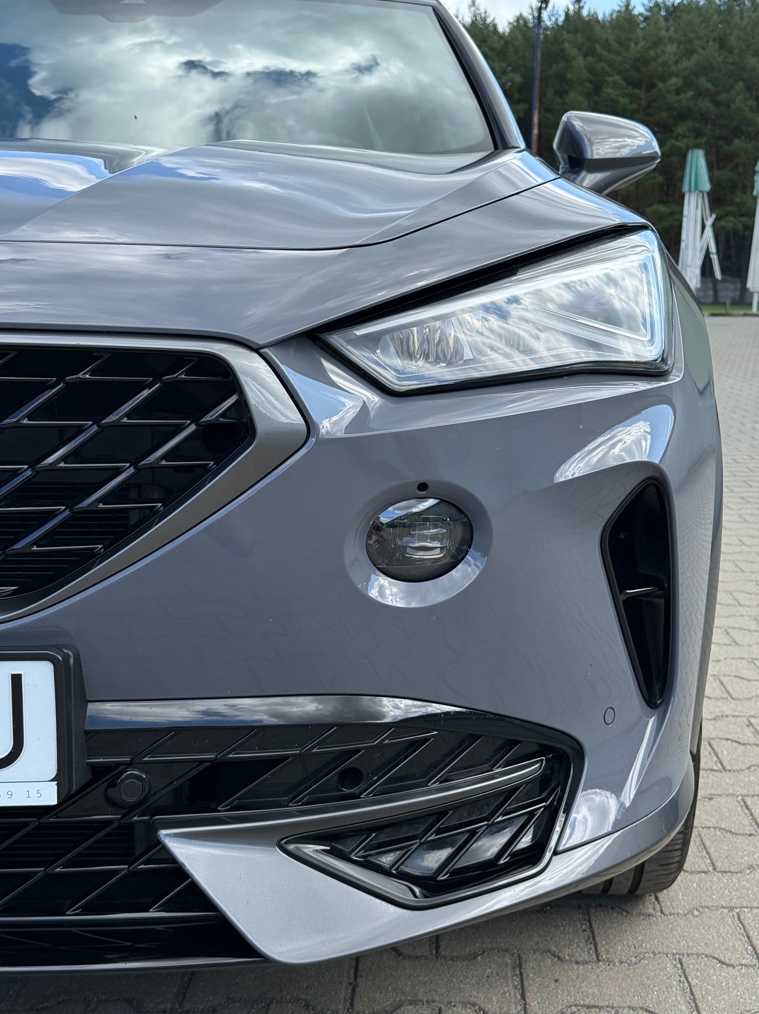 Szary samochód z reflektorem i kratką w zbliżeniu. Na masce odbija się niebo z chmurami. Widoczny fragment tablicy rejestracyjnej.