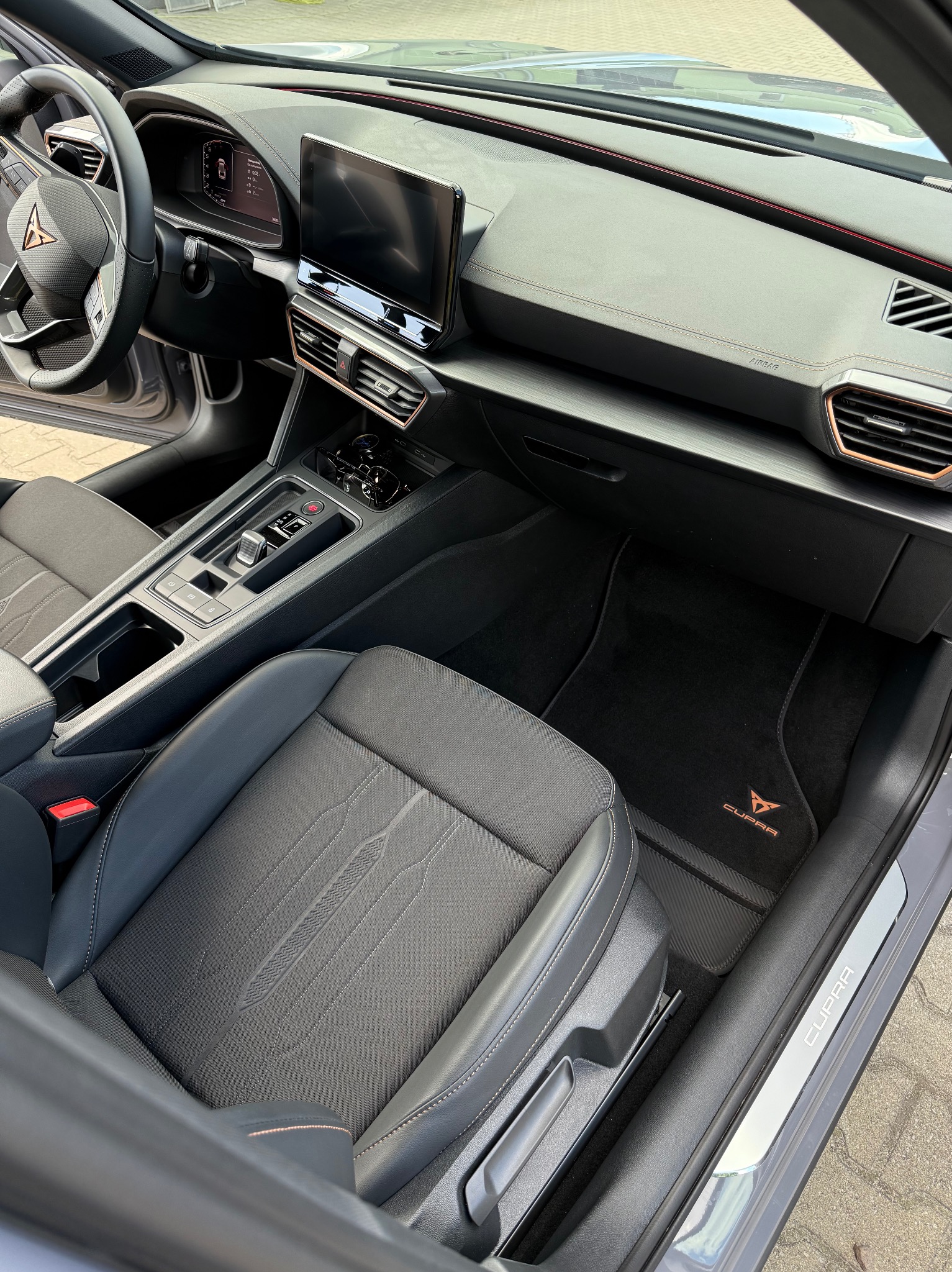 Wnętrze samochodu Cupra: czarna tapicerka fotela z subtelnym wzorem, deska rozdzielcza z ekranem dotykowym i detalami w kolorze miedzianym, eleganckie dywaniki z logo marki.