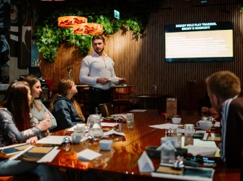 Prezentacja z ekranu w tle, mężczyzna gestykuluje, a wokół stołu siedzą słuchacze. Ciepłe oświetlenie, rośliny na ścianie tworzą przytulną atmosferę.