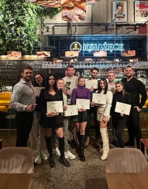 Grupa osób świętuje ukończenie kursu w barze, trzymając certyfikaty i trofeum. W tle bar z neonem 'Książęce' i rośliny. Oświetlenie ciepłe, tworzy przytulną atmosferę.