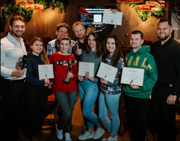 Grupa osób prezentuje certyfikaty i puchar po ukończonym szkoleniu w klimatycznym wnętrzu z drewnianą ścianą i roślinami. Widoczni mężczyźni i kobiety w różnym wieku.