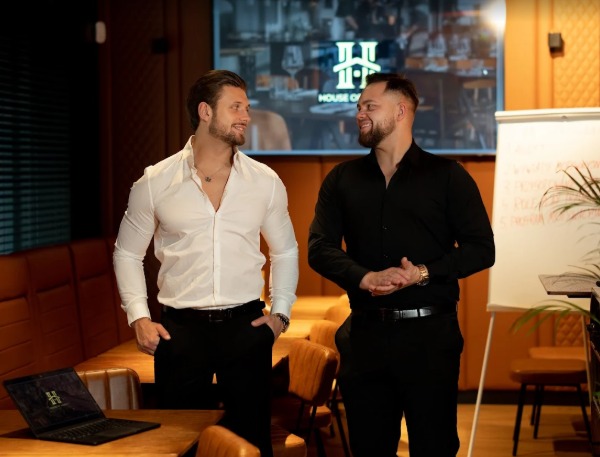 Dwóch uśmiechniętych mężczyzn w eleganckich koszulach w sali konferencyjnej z laptopem i tablicą. Branding firmy widoczny na laptopie i w tle.