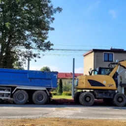 Ciężarówka i koparka CAT na tle budynku. Prace ziemne w toku. Niebieska ciężarówka z napisem 'Byens Største Natursteinsenter'.