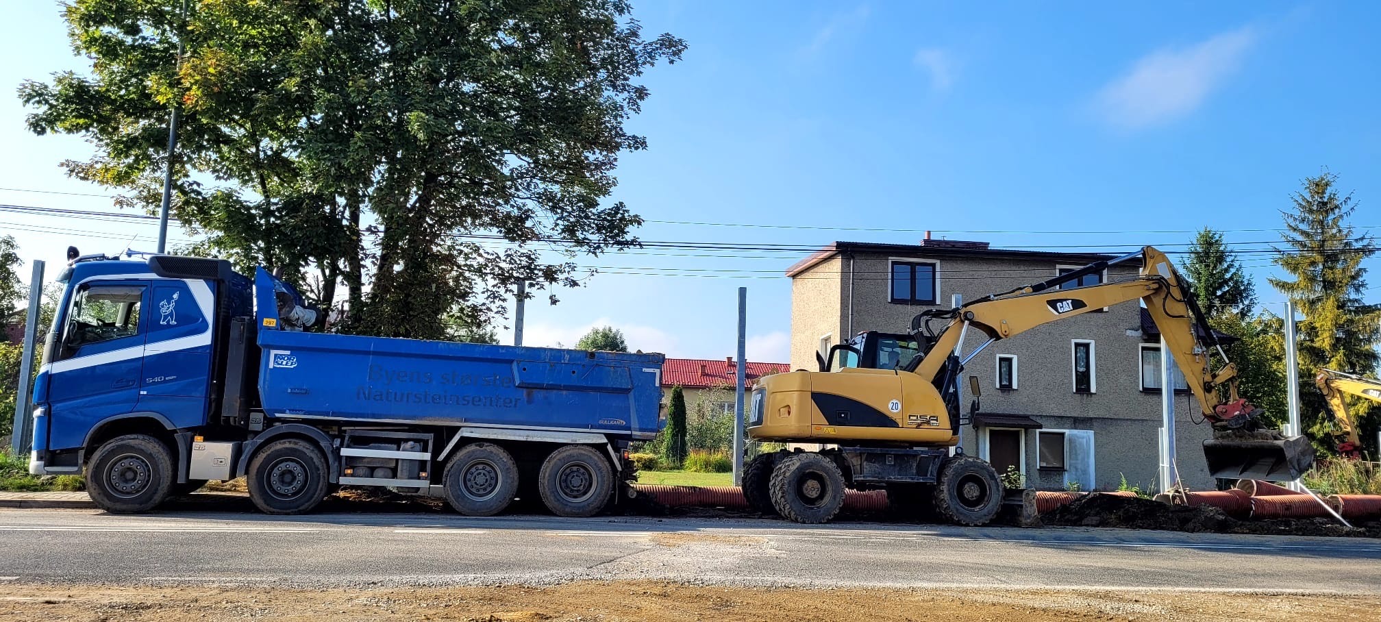 Ciężarówka i koparka CAT na tle budynku. Prace ziemne w toku. Niebieska ciężarówka z napisem 'Byens Største Natursteinsenter'.
