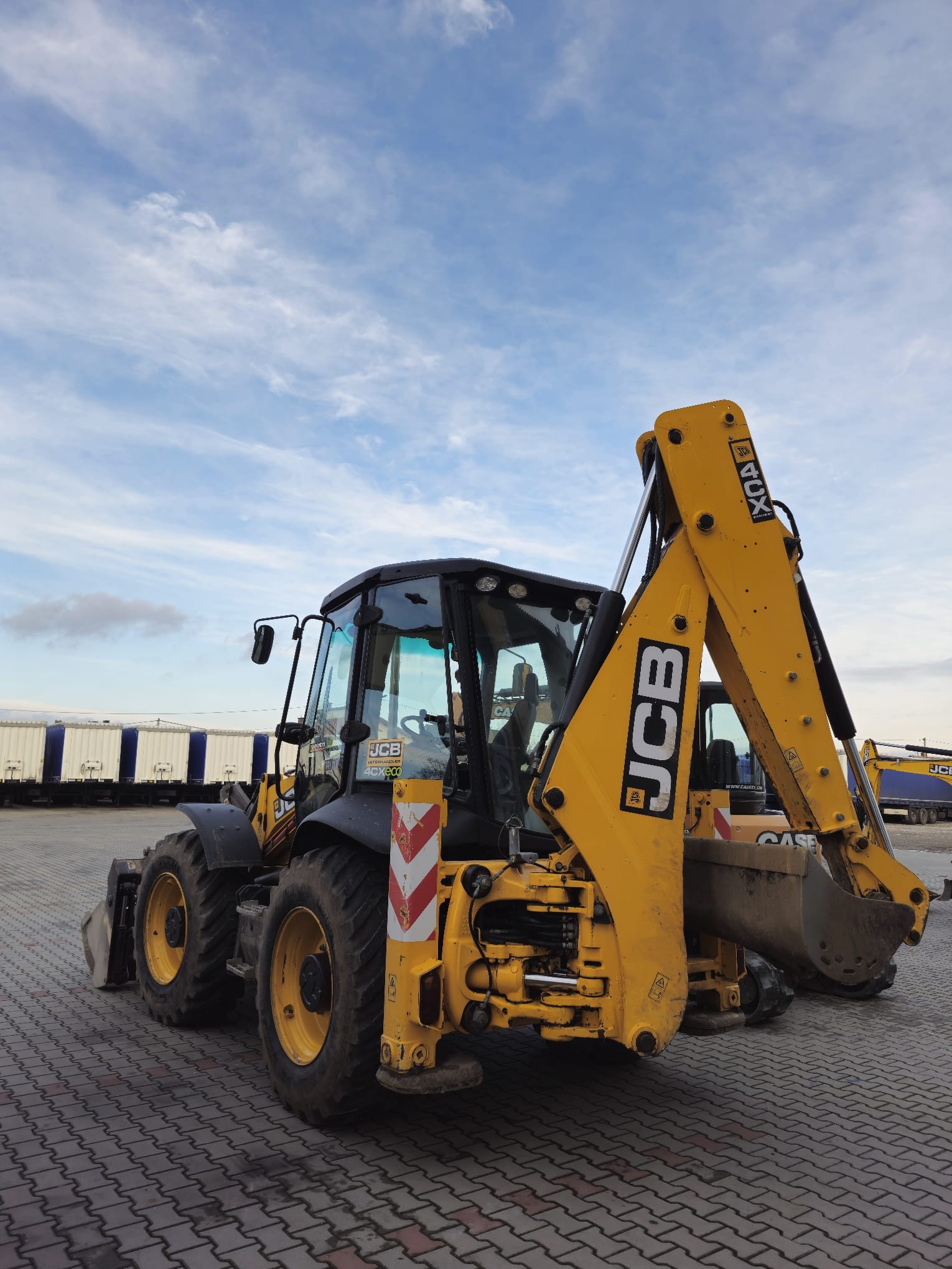Żółto-czarna koparko-ładowarka JCB na kostce brukowej, z wagonami w tle i błękitnym niebem. Widoczne oznaczenia ostrzegawcze i logo JCB. Maszyna gotowa do pracy.