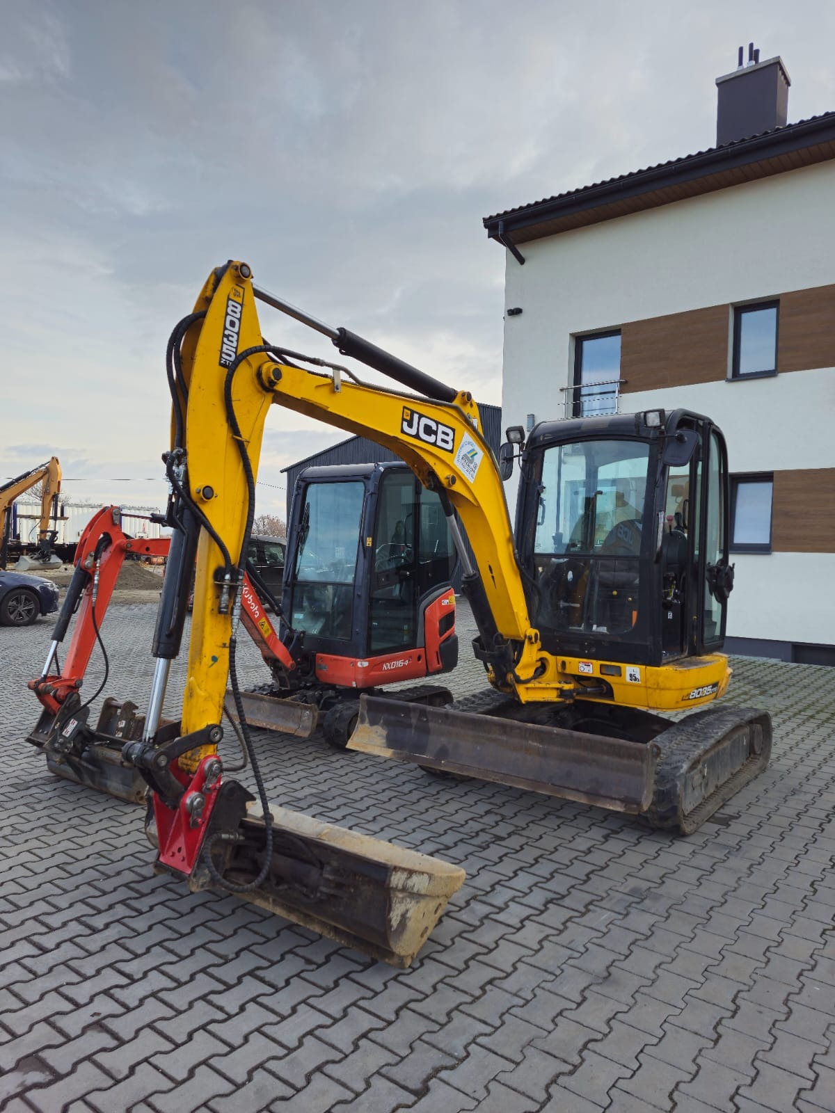 Dwa mini-koparki: żółta JCB 8035 i czerwona Kubota KX016-4 stoją obok siebie na brukowanej powierzchni, z budynkiem w tle.