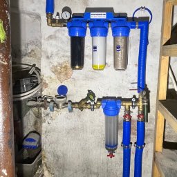 Pure Water Technology Jakub Szymczak - Złożony system filtrów wody z manometrem i niebieskimi rurami, zamontowany na ścianie z widocznymi śladami wilgoci. Obok drewniana drabina.