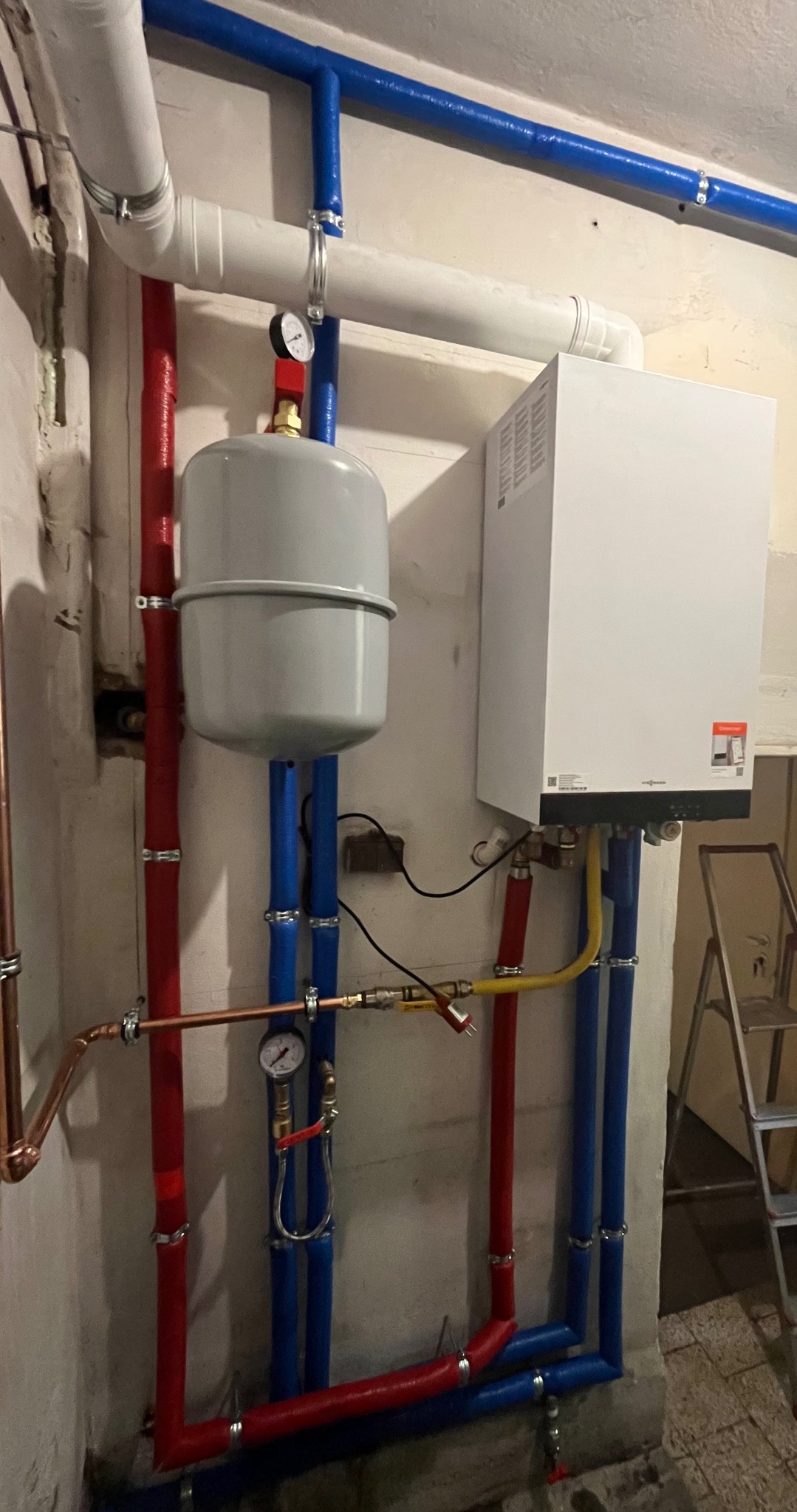 Biały piec CO zamontowany na ścianie, z szarym zbiornikiem wyrównawczym i kolorowymi rurami (czerwone, niebieskie, białe) w piwnicy. Widoczne manometry.