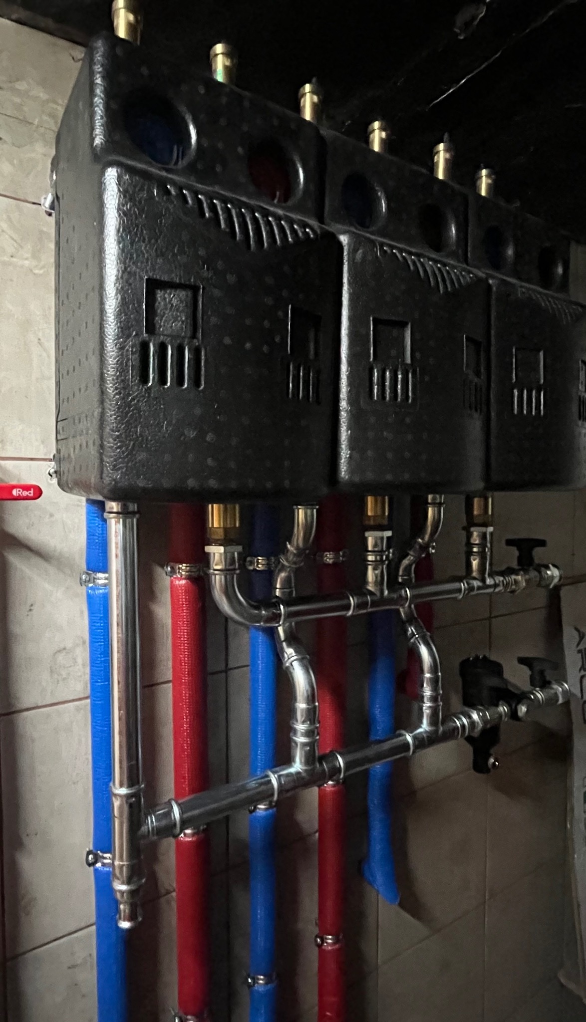 Czarny zbiornik z rzędami czerwonych i niebieskich rur, połączonych srebrnymi rurami z zaworami. Instalacja w pomieszczeniu z jasnymi płytkami.