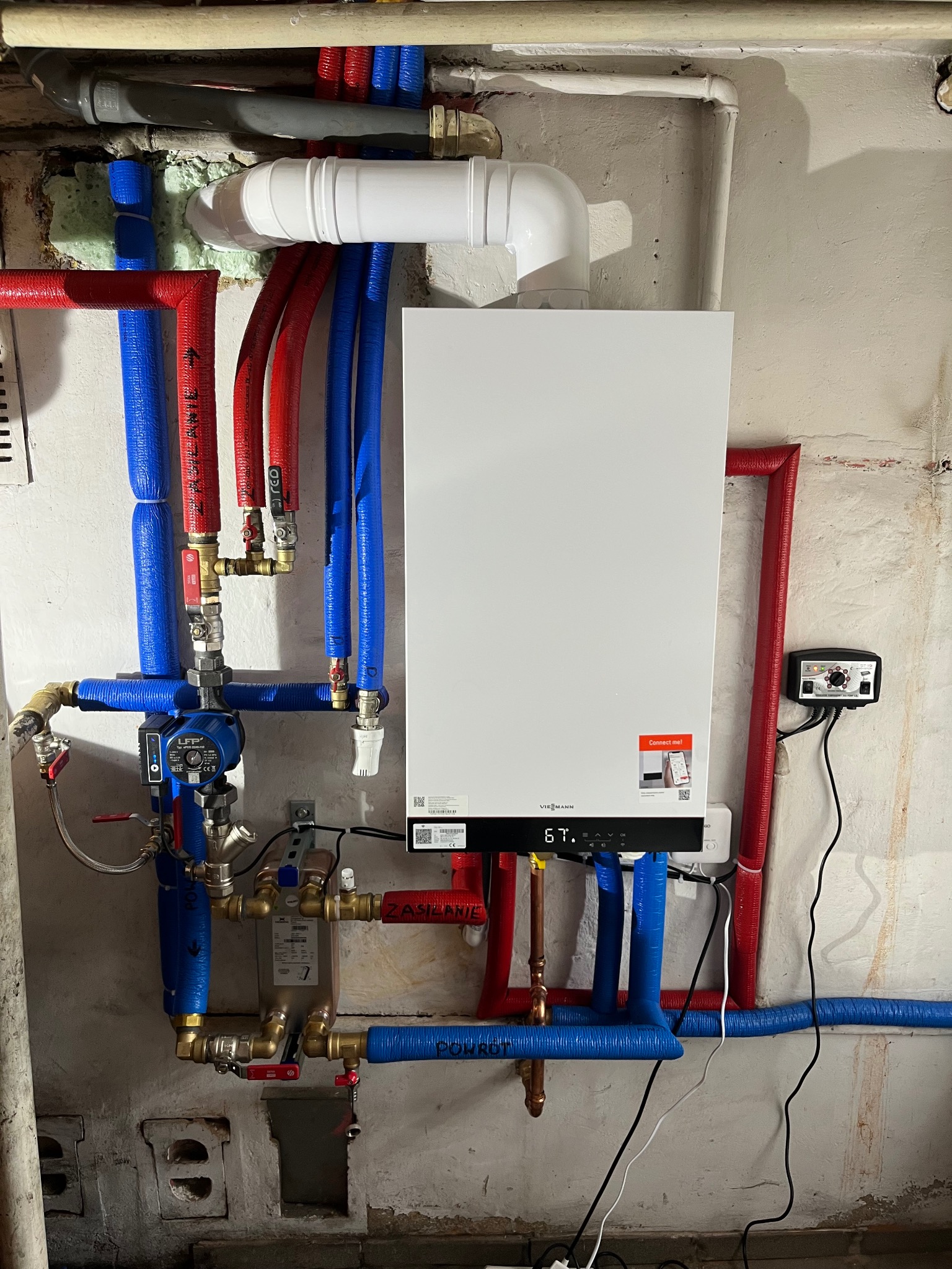 Nowoczesny, biały piec Viessmann z podłączonymi rurami CO w izolacji niebieskiej i czerwonej. Widoczne zawory, pompa obiegowa i sterownik. Tło: surowa, otynkowana ściana.