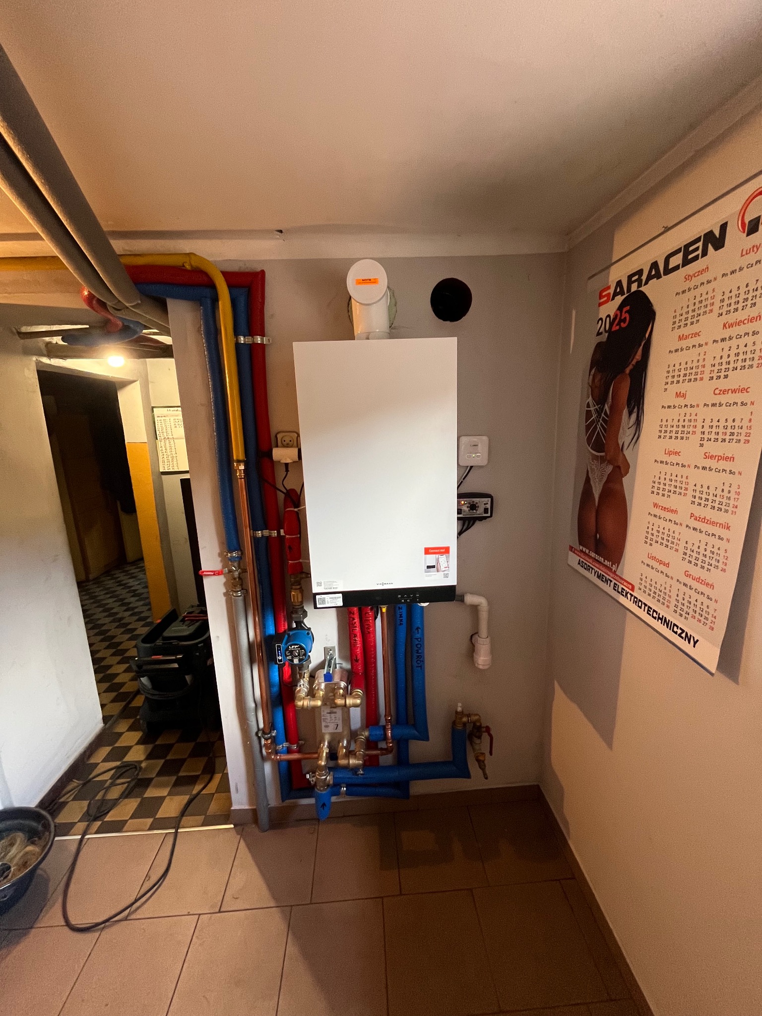 Nowoczesny piec gazowy Viessmann, zamontowany na ścianie, z podłączonymi rurami w kolorach niebieskim, czerwonym i żółtym. Widoczny układ zaworów i rur miedzianych.