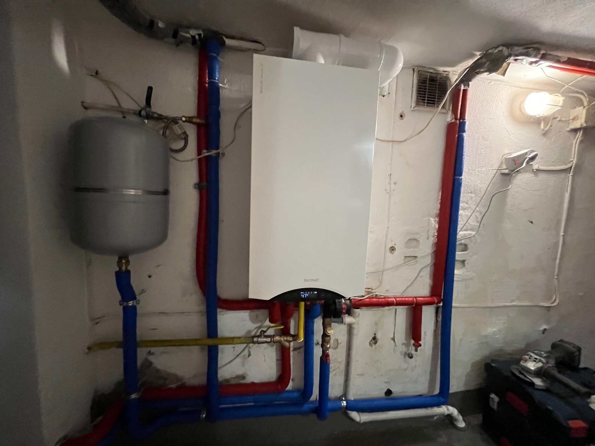 Biały piec gazowy marki Termet zamontowany na ścianie z widocznymi rurami w kolorach niebieskim i czerwonym. Obok szary zbiornik wyrównawczy i narzędzia.