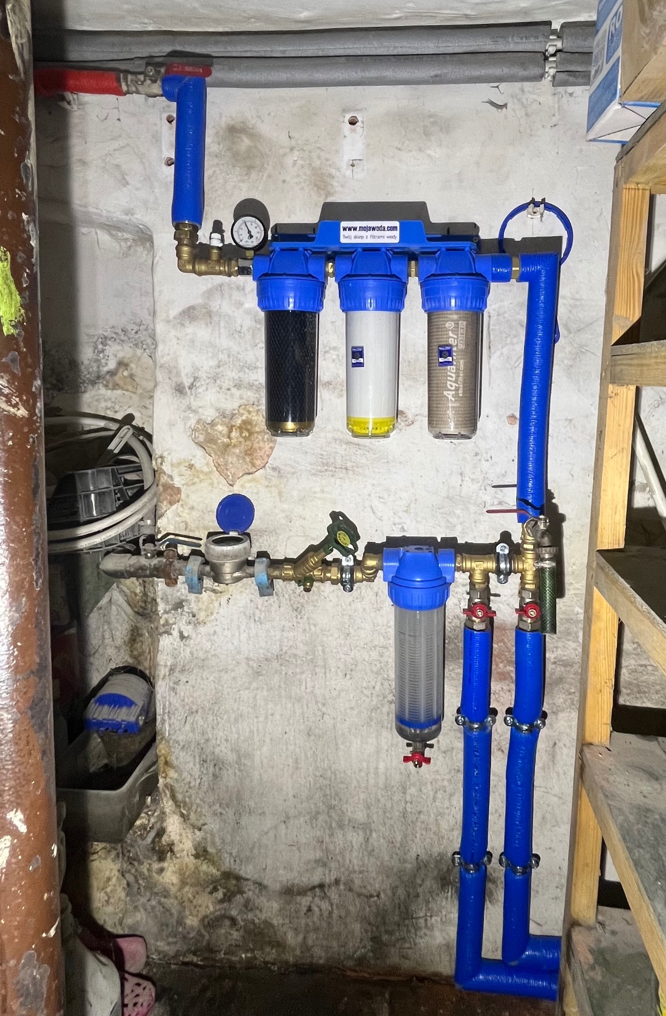 Złożony system filtrów wody z manometrem i niebieskimi rurami, zamontowany na ścianie z widocznymi śladami wilgoci. Obok drewniana drabina.