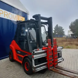 Czerwono-czarny wózek widłowy Fenwick H60 BG z osprzętem, zaparkowany na zewnątrz obok niebieskiej hali z napisem 'PROMLIFT' w pochmurny dzień.