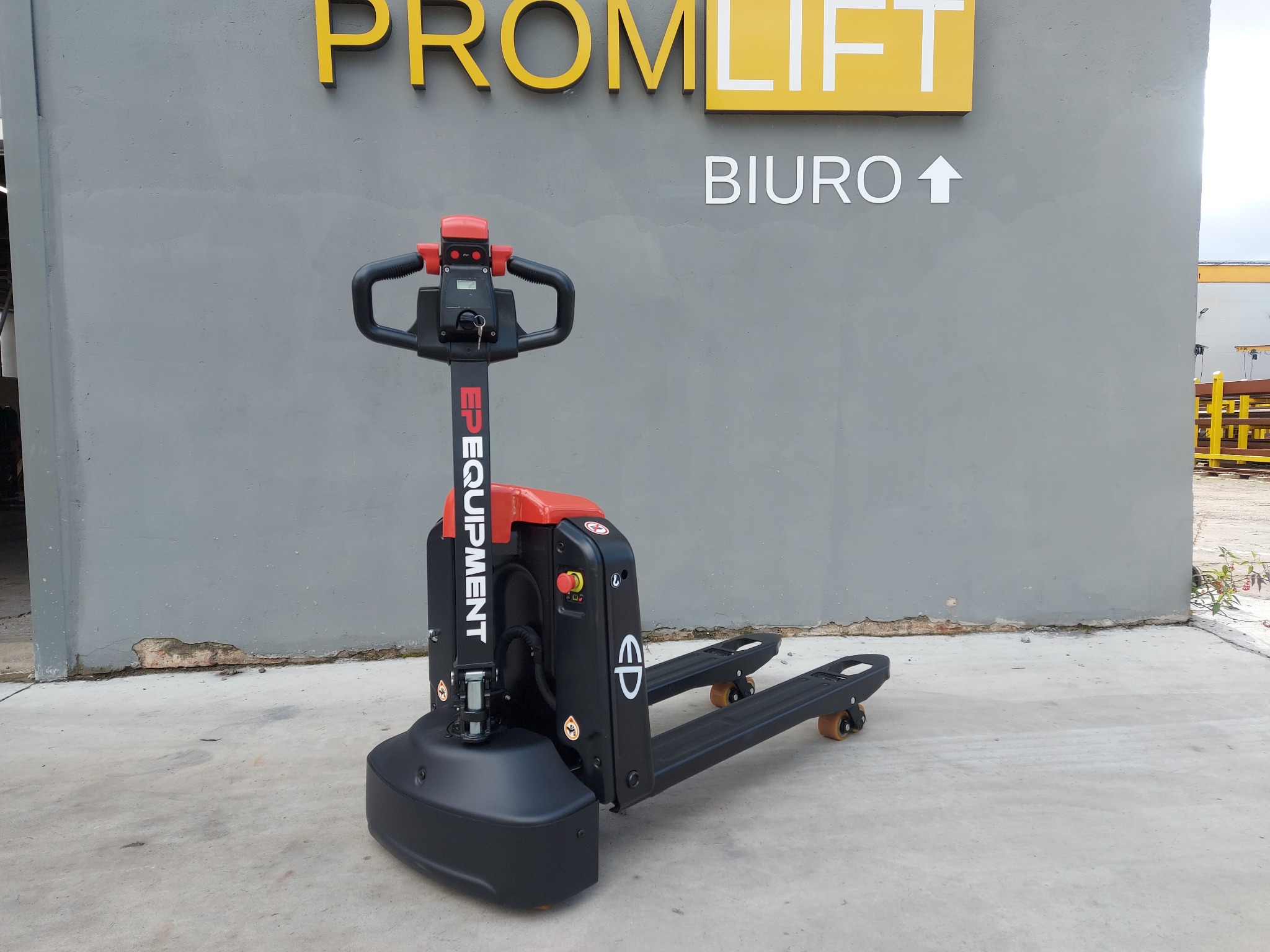 Elektryczny wózek paletowy EP Equipment na tle szarej ściany z logo firmy Promlift i strzałką wskazującą biuro. Wózek czarno-czerwony, widoczny z boku.