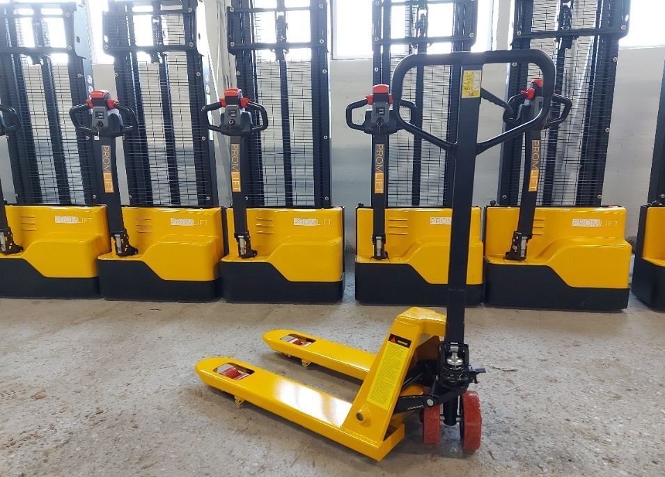 Żółty wózek paletowy ręczny Promlift na betonowej podłodze, z rzędem wózków paletowych elektrycznych w tle. Widok z przodu, lekko z dołu.