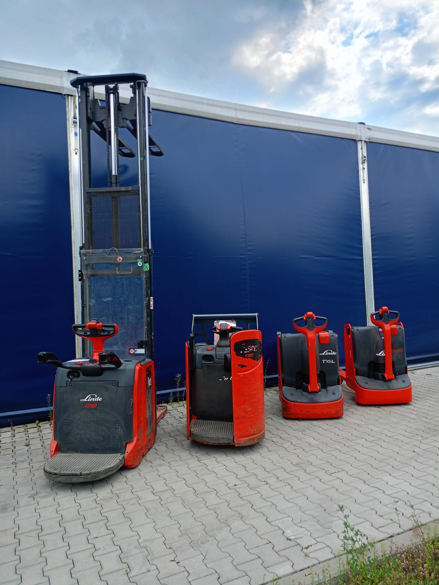Cztery czerwono-czarne wózki paletowe Linde na kostce brukowej przed niebieskim namiotem. Jeden z wózków to model D12 z wysokim masztem.