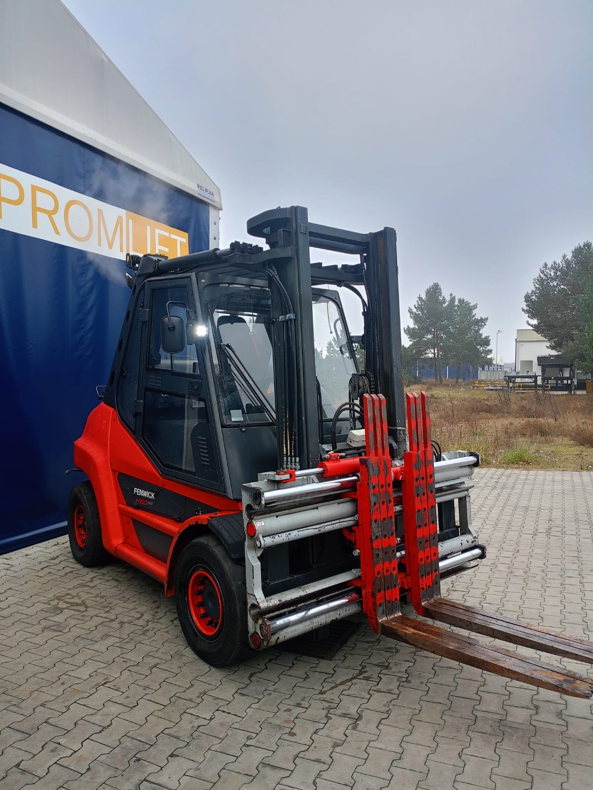 Czerwono-czarny wózek widłowy Fenwick H60 BG z osprzętem, zaparkowany na zewnątrz obok niebieskiej hali z napisem 'PROMLIFT' w pochmurny dzień.