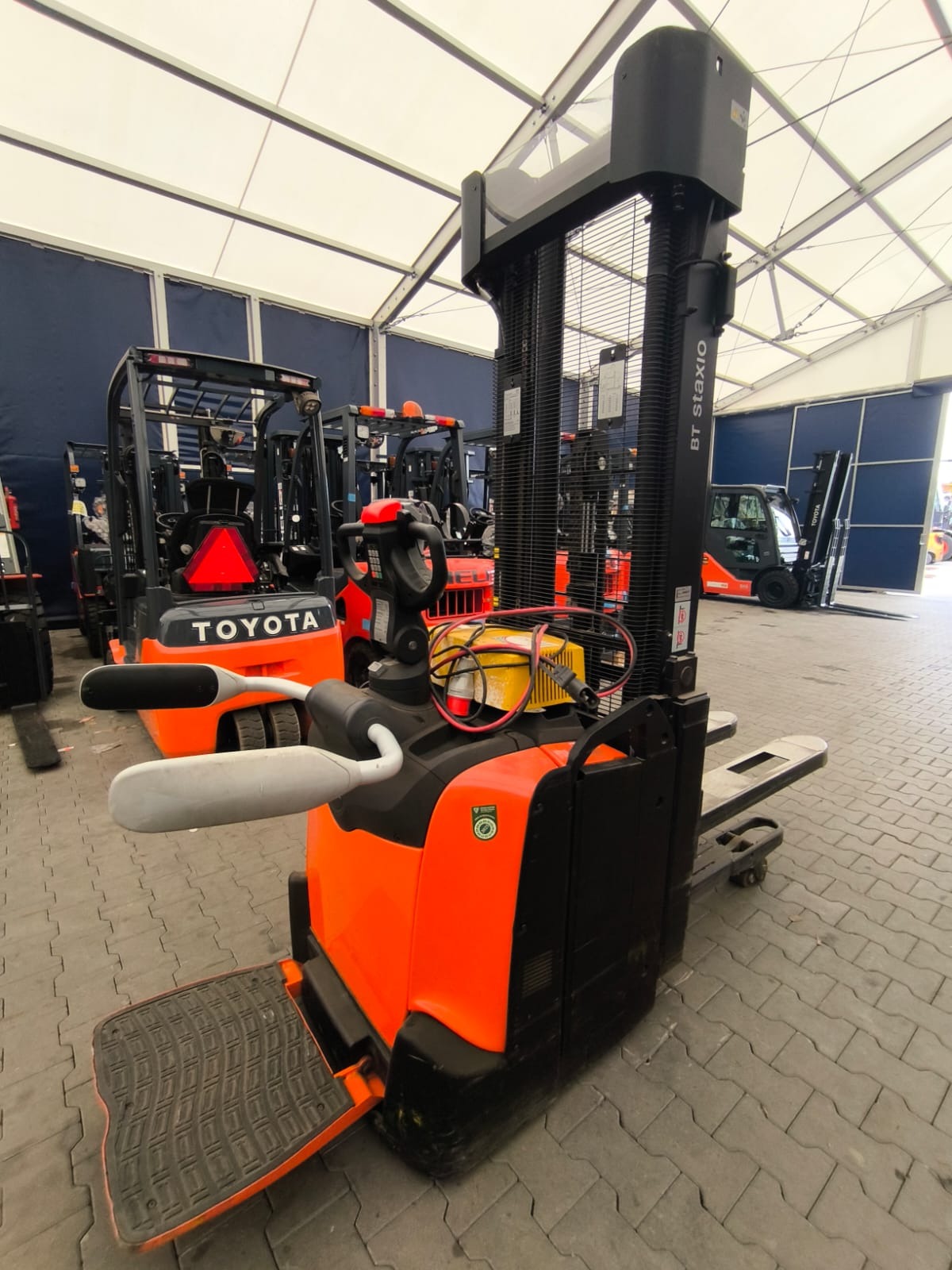 Pomarańczowo-czarny wózek paletowy elektryczny BT Staxio na tle innych wózków widłowych Toyota w hali z białym sufitem i niebieskimi ścianami.