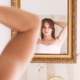 PRO-INVEST - Panna młoda w odbiciu lustra w złotej ramie, poprawia włosy. W tle beżowe płytki i złota bateria umywalkowa. Część ręki w lewym dolnym rogu zdjęcia.
