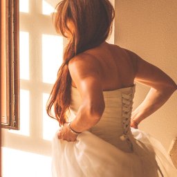 PRO-INVEST - Panna młoda w sukni ślubnej, z plecami w gorsecie i rozpuszczonymi włosami, stoi w ciepłym świetle okna z kratownicą. Ujęcie od tyłu, delikatne oświetlenie.