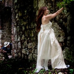 PRO-INVEST - Panna młoda w sukni ślubnej i trampkach pozuje na tle kamiennej ściany, w tle mężczyzna z psem przy ceglanym łuku. Nietypowe ujęcie, oryginalna sesja.