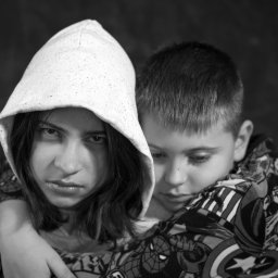 PRO-INVEST - Czarno-białe portretowe ujęcie dwójki dzieci, z dziewczynką w kapturze na pierwszym planie i chłopcem w bluzie z motywami komiksowymi w tle, wyrażające emocje.