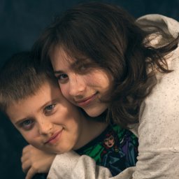 PRO-INVEST - Portret rodzeństwa: uśmiechnięty chłopiec z niebieskimi oczami obejmowany przez starszą siostrę w białej bluzie z kapturem na ciemnym tle. Widoczny rysunek na koszulce chłopca.