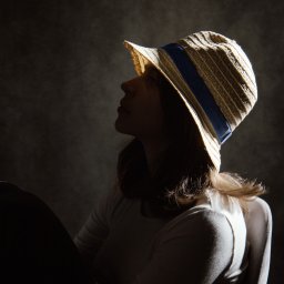 PRO-INVEST - Portret kobiety w słomkowym kapeluszu z niebieską wstążką, oświetlonej z góry, z profilu na ciemnym tle. Ujęcie w klimacie retro.