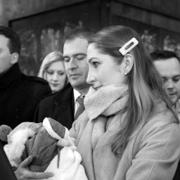 PRO-INVEST - Czarno-białe ujęcie z uroczystości chrztu: kobieta z ozdobną spinką we włosach trzyma dziecko, w tle widoczni goście i fragment obrazu na ścianie kościoła.