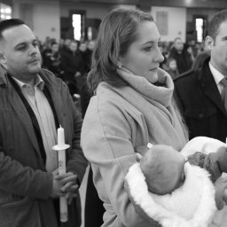 PRO-INVEST - Czarno-białe zdjęcie z ceremonii chrztu: kobieta trzyma niemowlę, obok mężczyzna z świecą, w tle zgromadzeni ludzie w kościele.