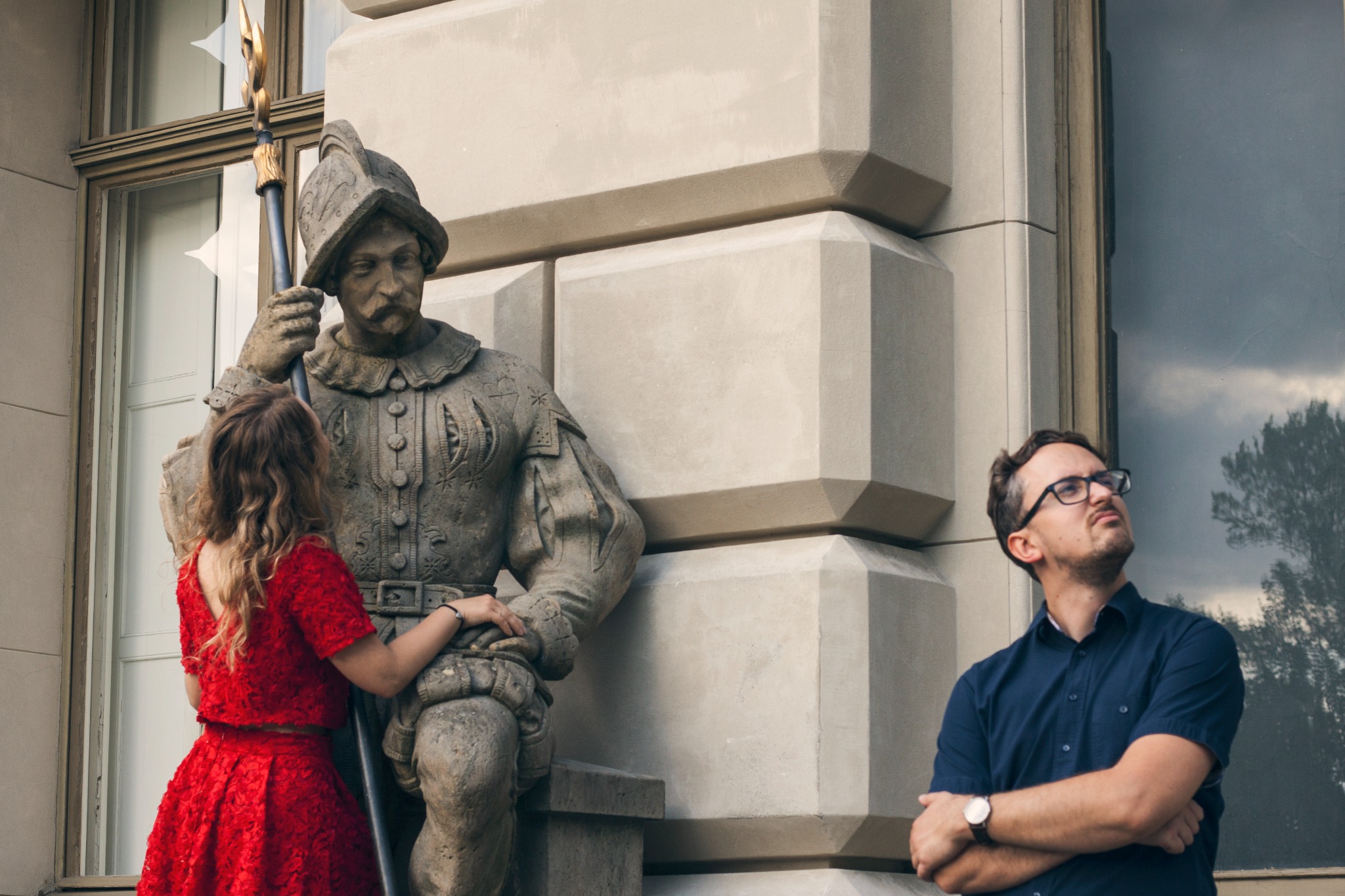Sesja zdjęciowa z rzeźbą: Kobieta w czerwonej sukience opiera się o kamienną figurę żołnierza, mężczyzna w okularach patrzy w górę, jasne tło budynku.