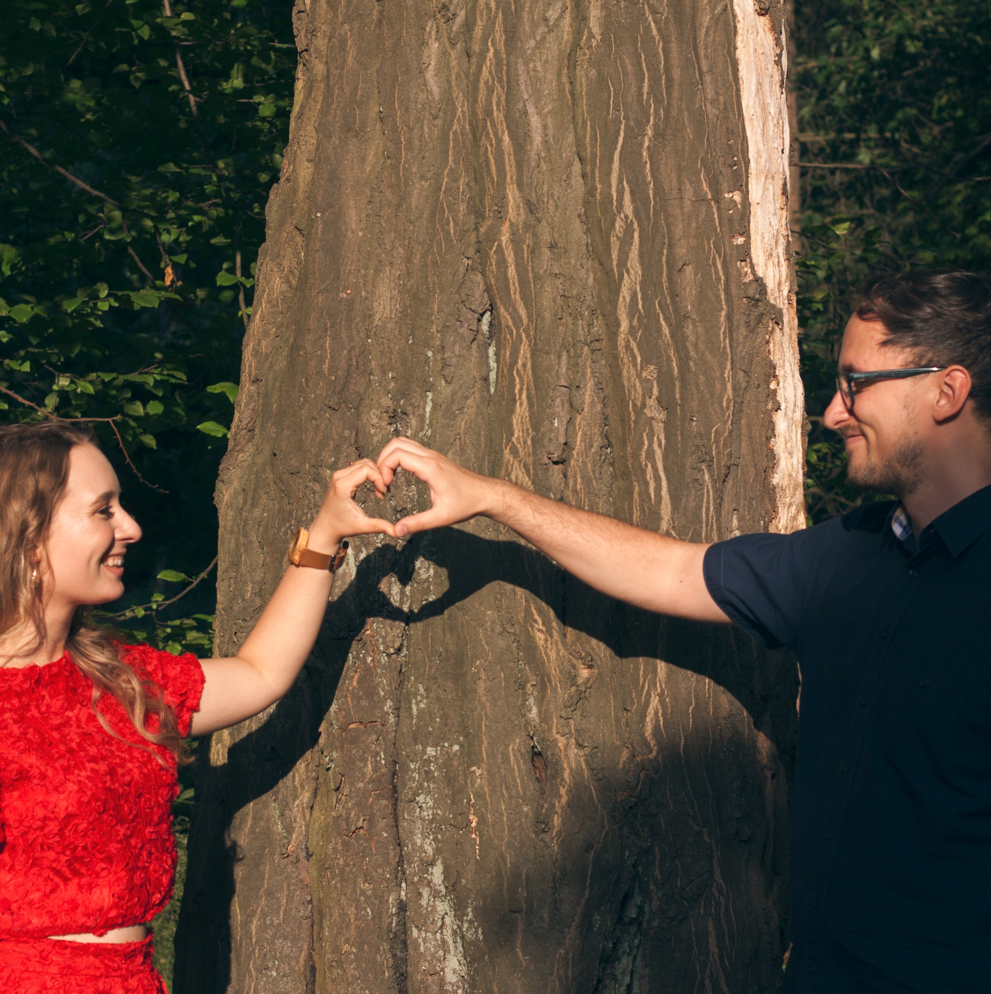 Zakochana para w plenerze tworzy dłońmi cień w kształcie serca na korze drzewa. Uśmiechnięci, patrzą na siebie. Ona w czerwonej sukience, on w granatowej koszuli.