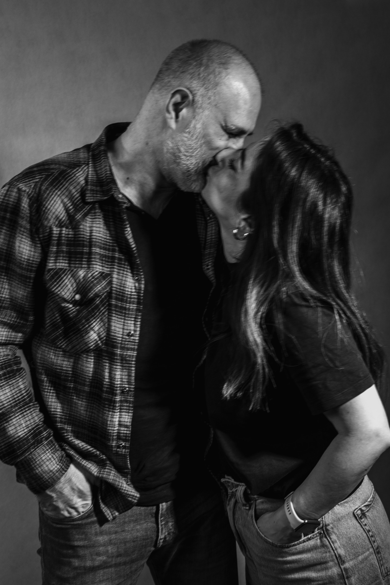 Czarno-białe zdjęcie pary w czułym pocałunku, on w koszuli w kratę, ona z kolczykiem kołem. Oboje w jeansach z rękami w kieszeniach, na ciemnym tle. Intymna sesja.