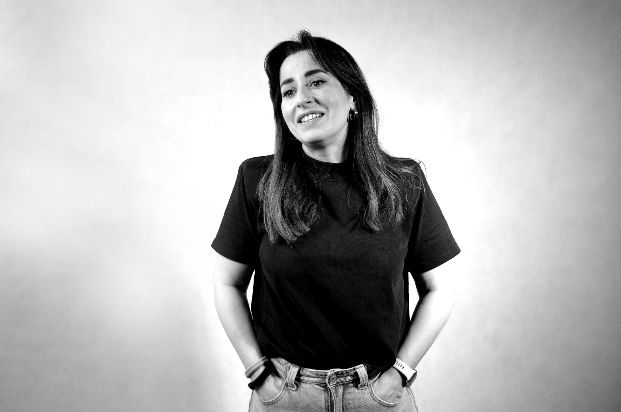 Czarno-białe zdjęcie uśmiechniętej kobiety w dżinsach i czarnym T-shircie, z rękami w kieszeniach, na jasnym tle. Portret w naturalnym oświetleniu.