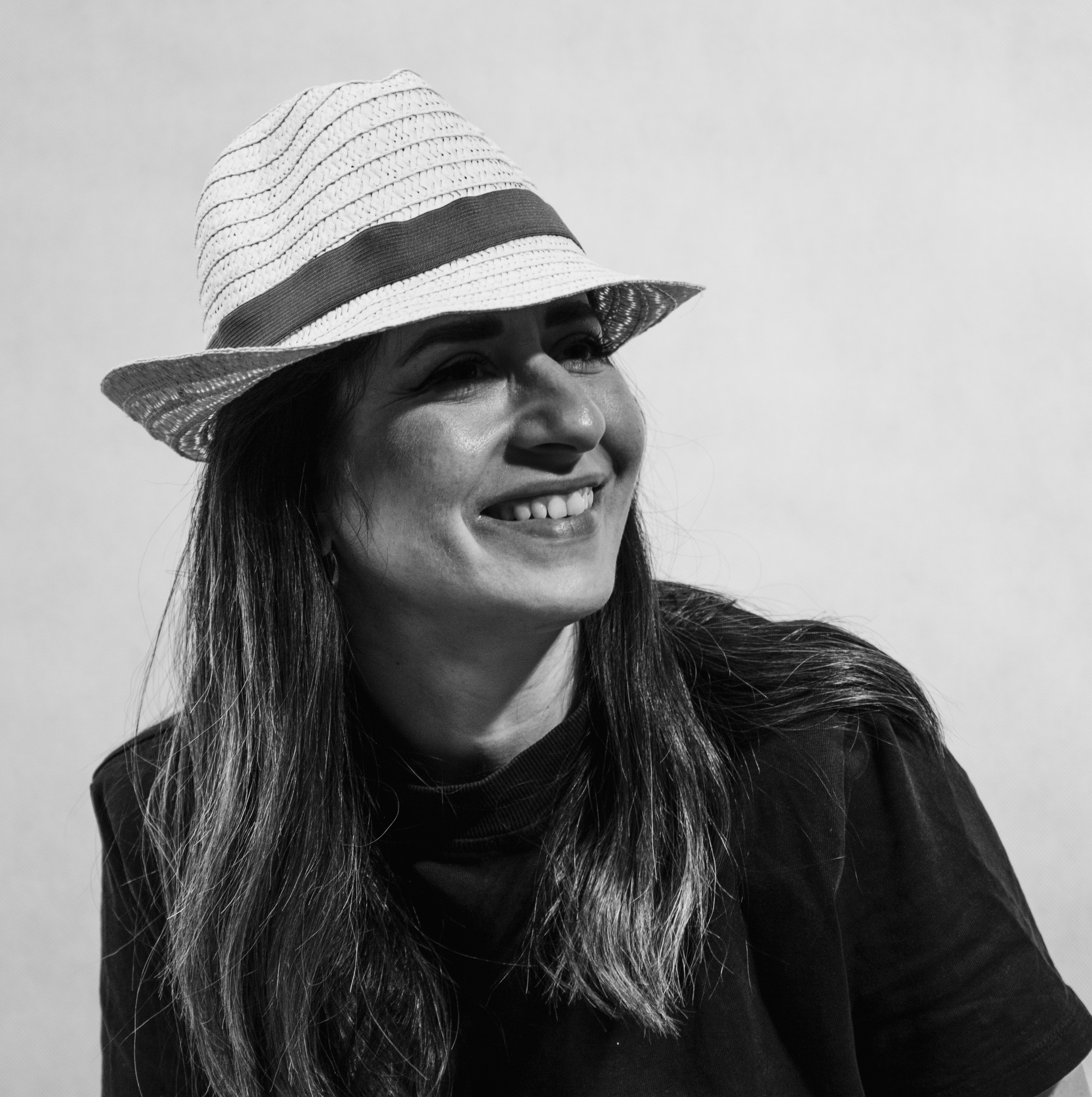 Czarno-białe, uśmiechnięte portretowe ujęcie kobiety w słomkowym kapeluszu, z długimi włosami i ciemnej bluzce na jasnym tle. Patrzy w prawo, oświetlona z lewej strony.