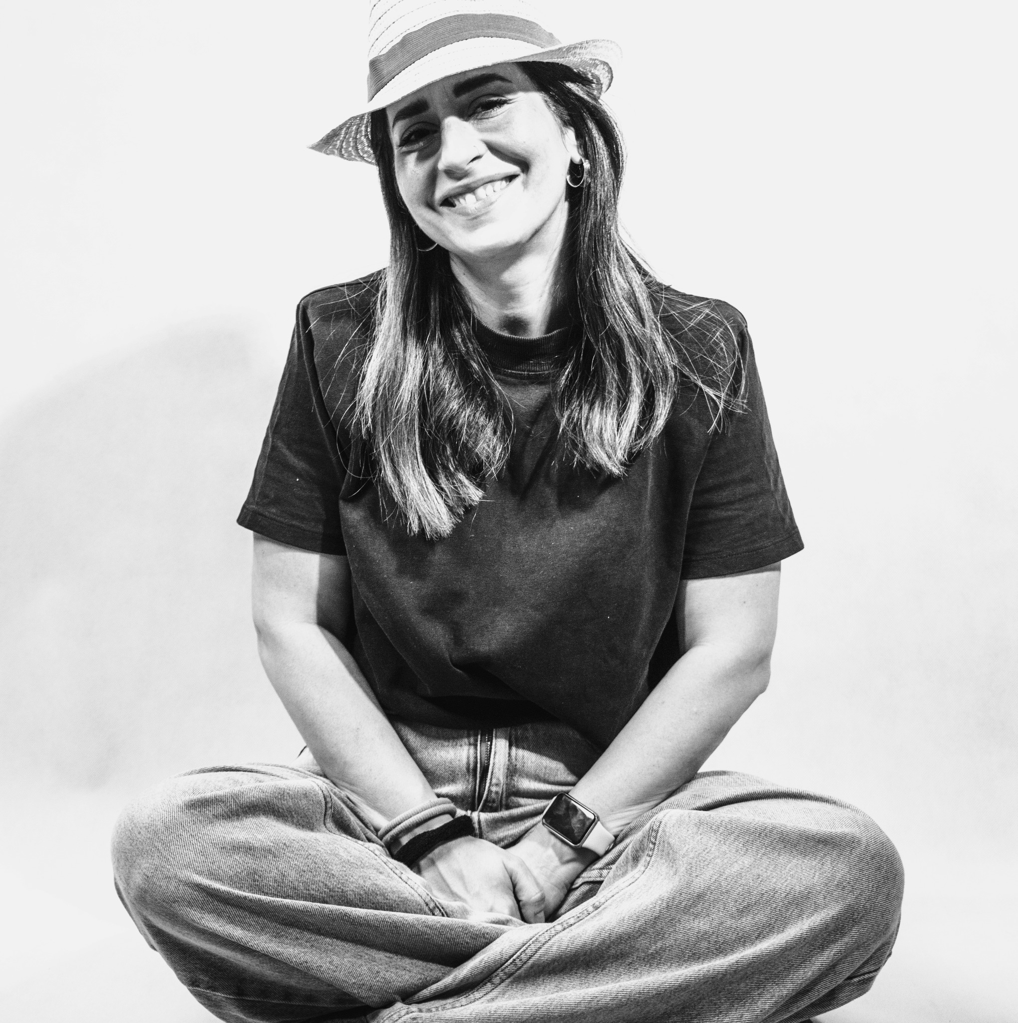 Portret uśmiechniętej kobiety w kapeluszu, siedzącej po turecku na jasnym tle. Zdjęcie czarno-białe, z widocznymi detalami stroju i akcesoriów.