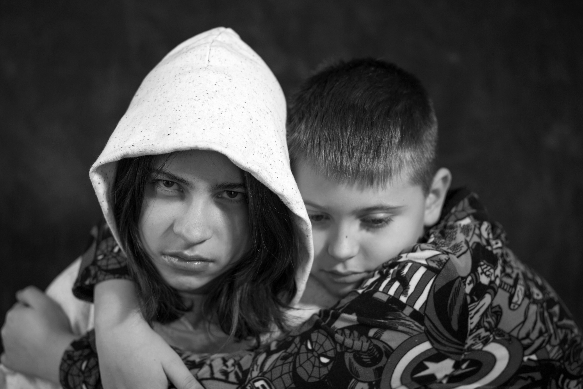 Czarno-białe portretowe ujęcie dwójki dzieci, z dziewczynką w kapturze na pierwszym planie i chłopcem w bluzie z motywami komiksowymi w tle, wyrażające emocje.