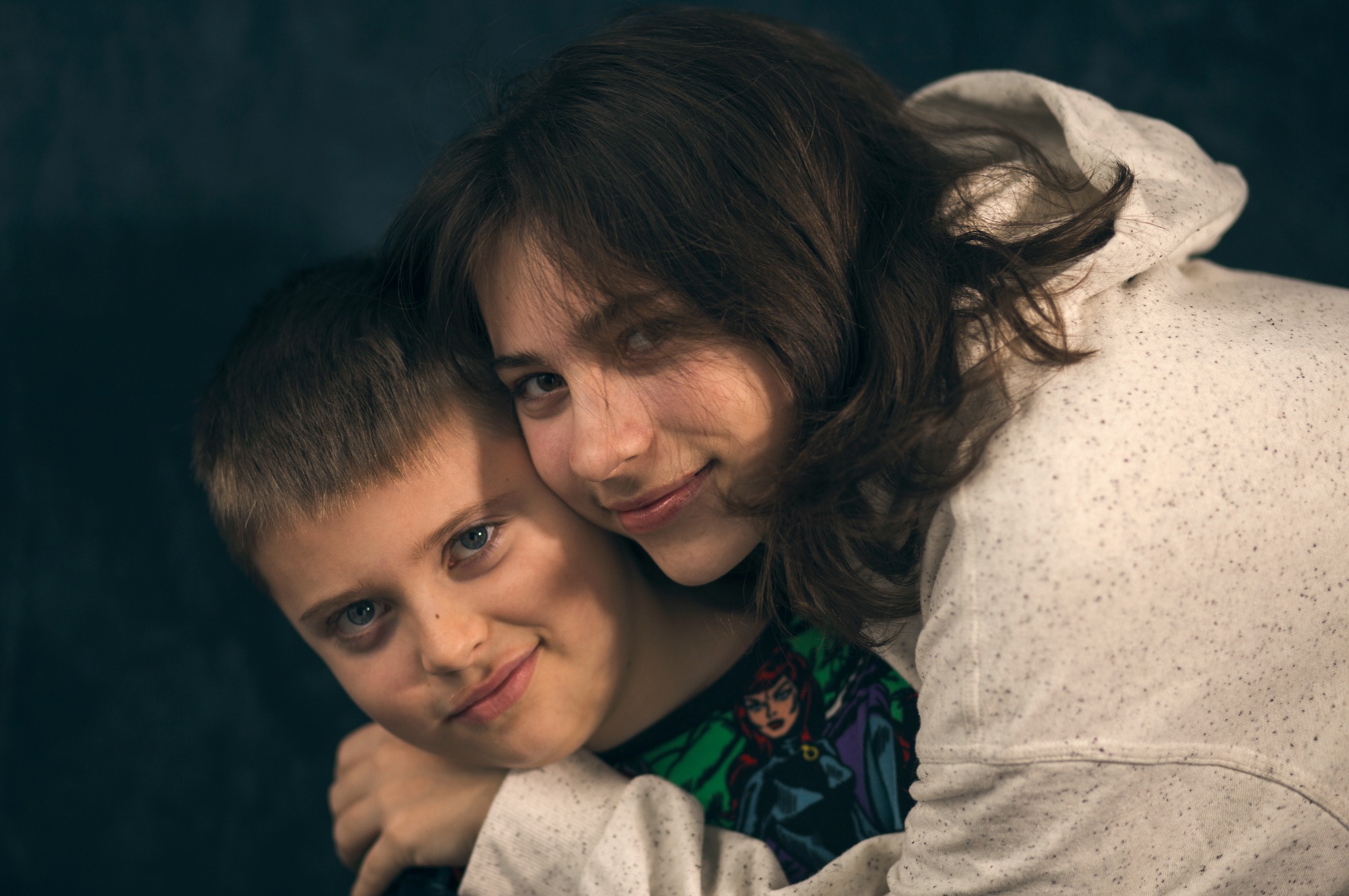 Portret rodzeństwa: uśmiechnięty chłopiec z niebieskimi oczami obejmowany przez starszą siostrę w białej bluzie z kapturem na ciemnym tle. Widoczny rysunek na koszulce chłopca.