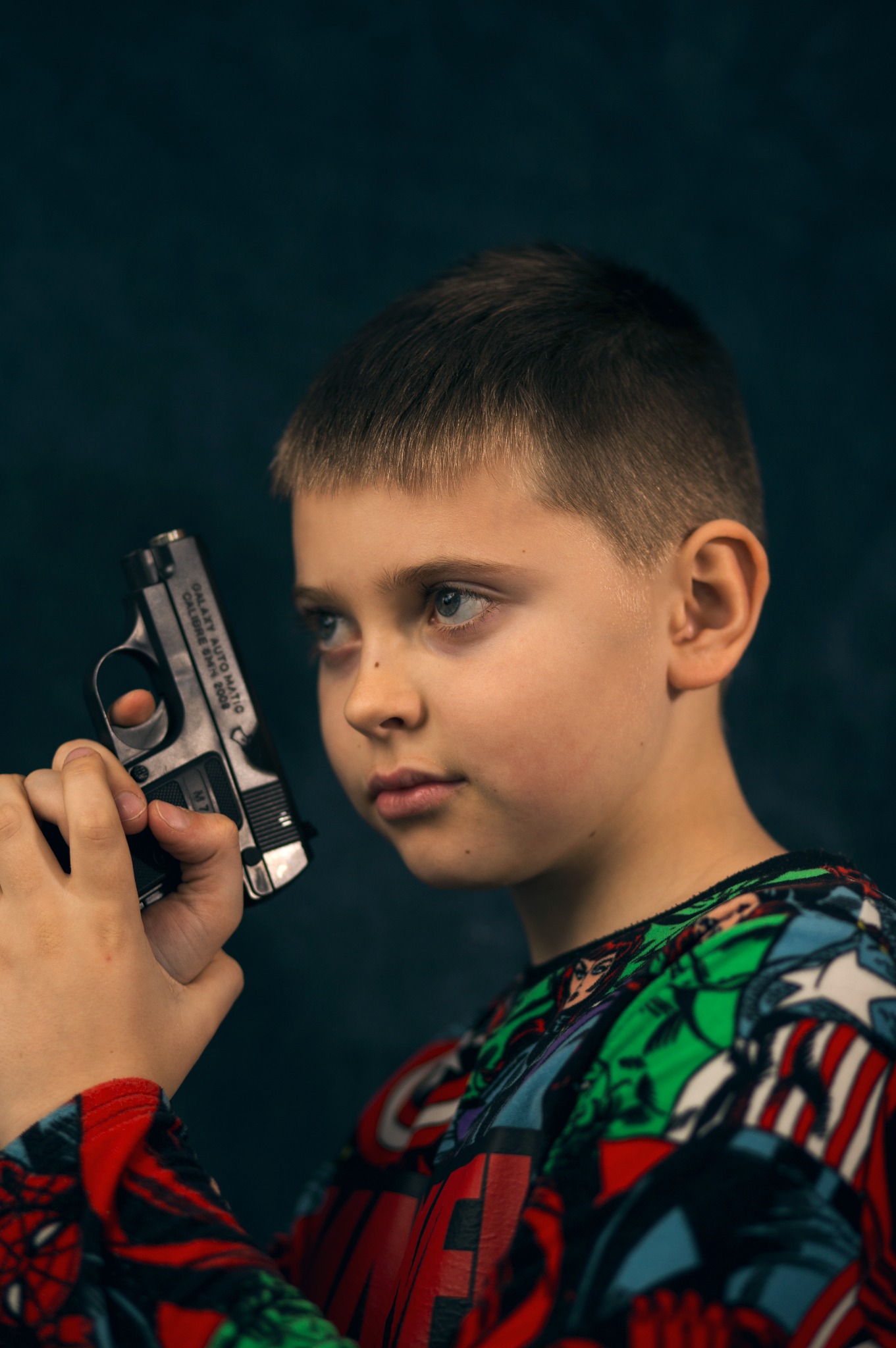 Portret chłopca trzymającego pistolet zabawkę, ubrany w bluzę z motywami superbohaterów na ciemnym tle. Ujęcie z profilu, skupione na twarzy i dłoniach.
