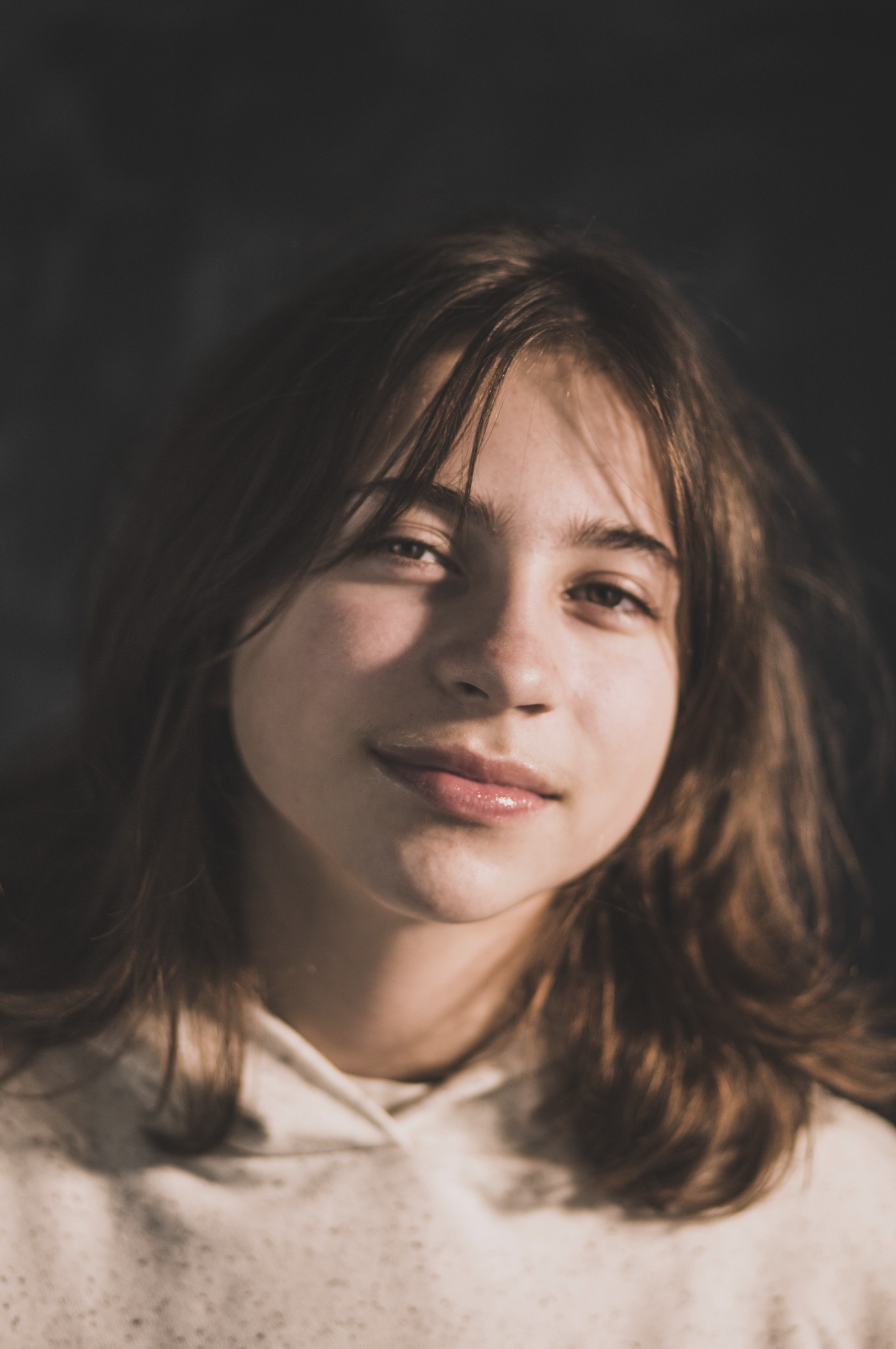 Portret młodej kobiety z długimi, brązowymi włosami i delikatnym uśmiechem, oświetlonej naturalnym światłem na ciemnym tle. Ujęcie z bliska, skupiające się na wyrazie twarzy.