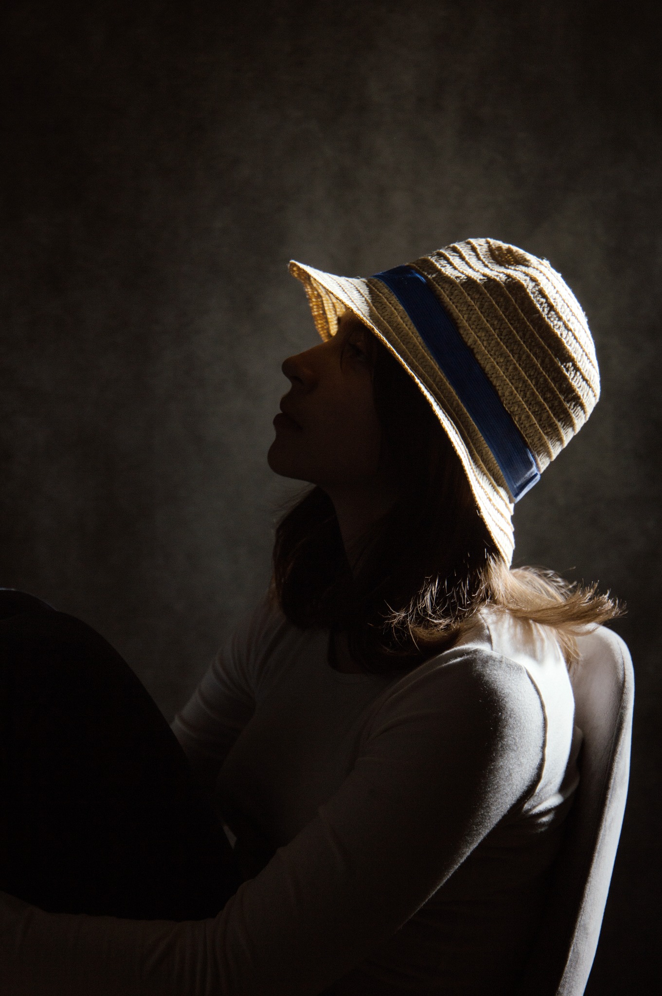 Portret kobiety w słomkowym kapeluszu z niebieską wstążką, oświetlonej z góry, z profilu na ciemnym tle. Ujęcie w klimacie retro.